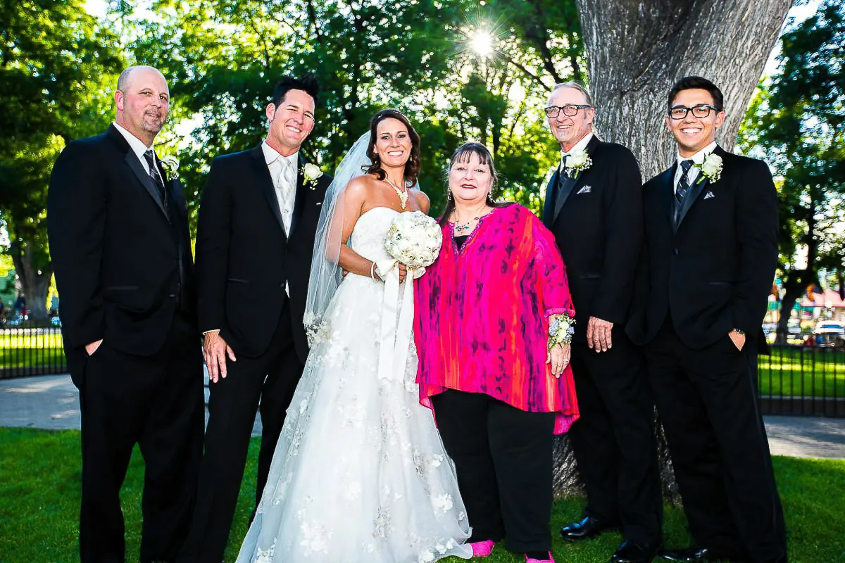 Wedding Party Photos A bride and groom pose for wedding photography with four others in a park. The bride, in a white gown with a bouquet, stands beside the groom and three men in black suits. A woman in bright pink takes center stage. All are smiling, framed by trees and sunlight.