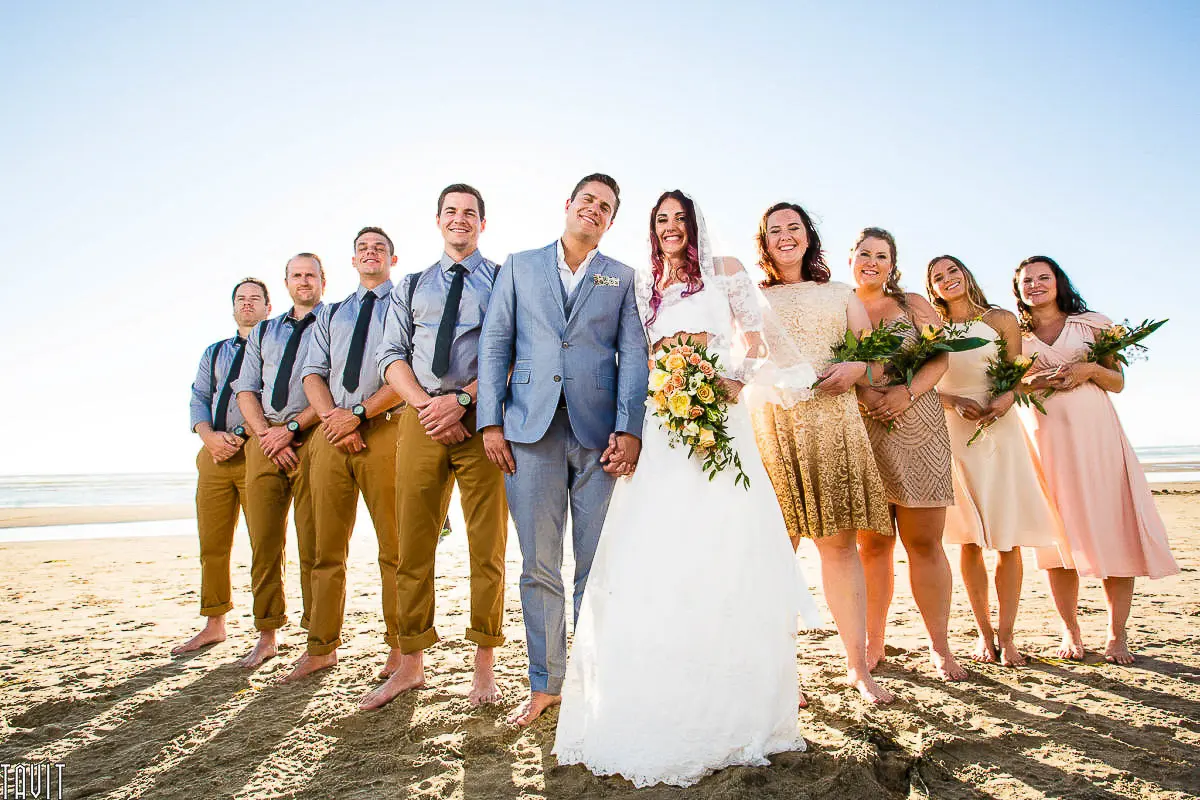 Wedding Party Photos A bride and groom stand barefoot on a sunny beach, perfectly captured in wedding photography. The groom is in a light blue suit, the bride radiant in her white dress with a bouquet. Bridesmaids and groomsmen, dressed informally, smile brightly while holding flowers.