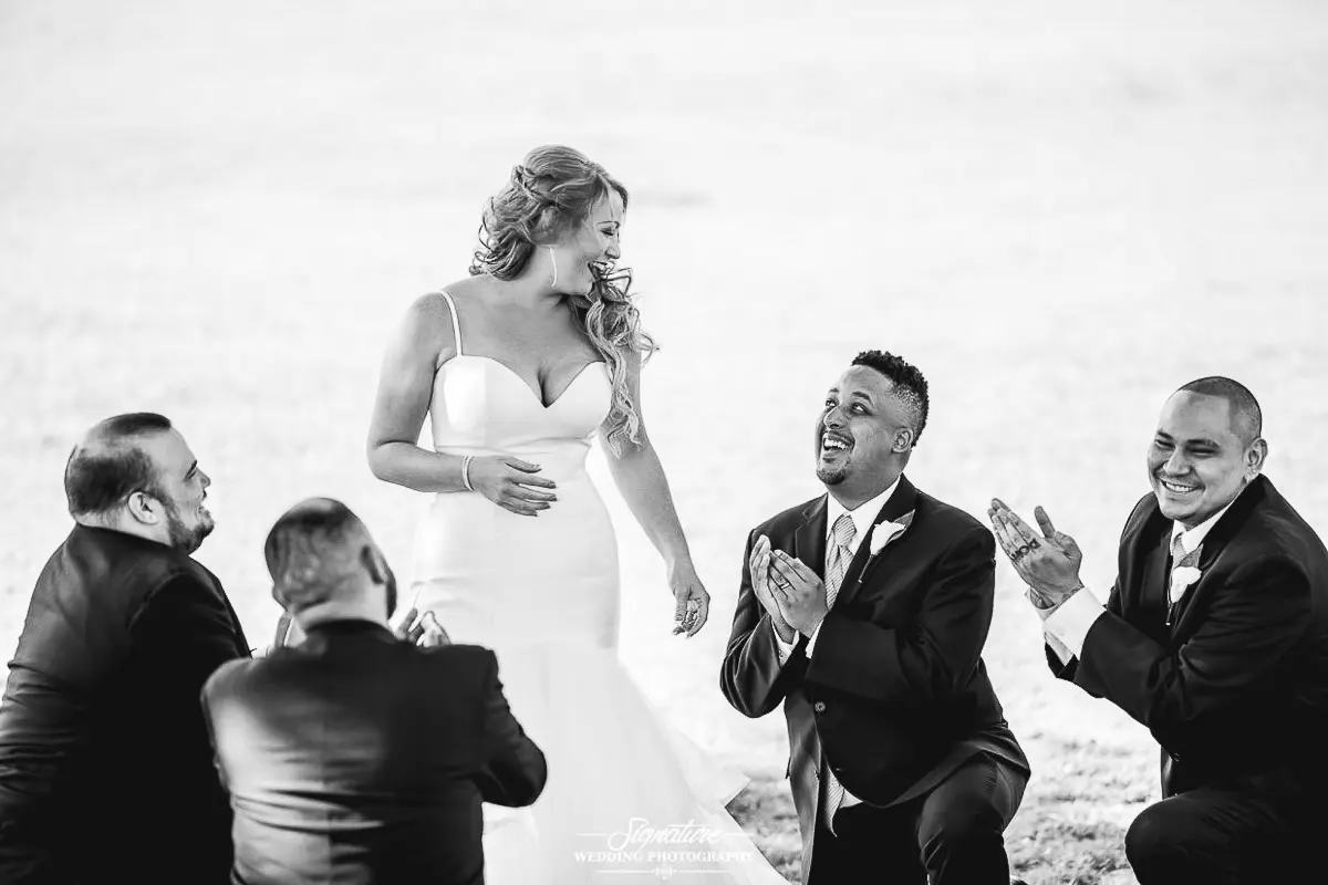 Wedding Party Photos A bride in a gown stands smiling as four groomsmen, dressed in suits, kneel around her and gesture playfully. The wedding photography captures this joyful moment in timeless black and white against a serene grassy backdrop.