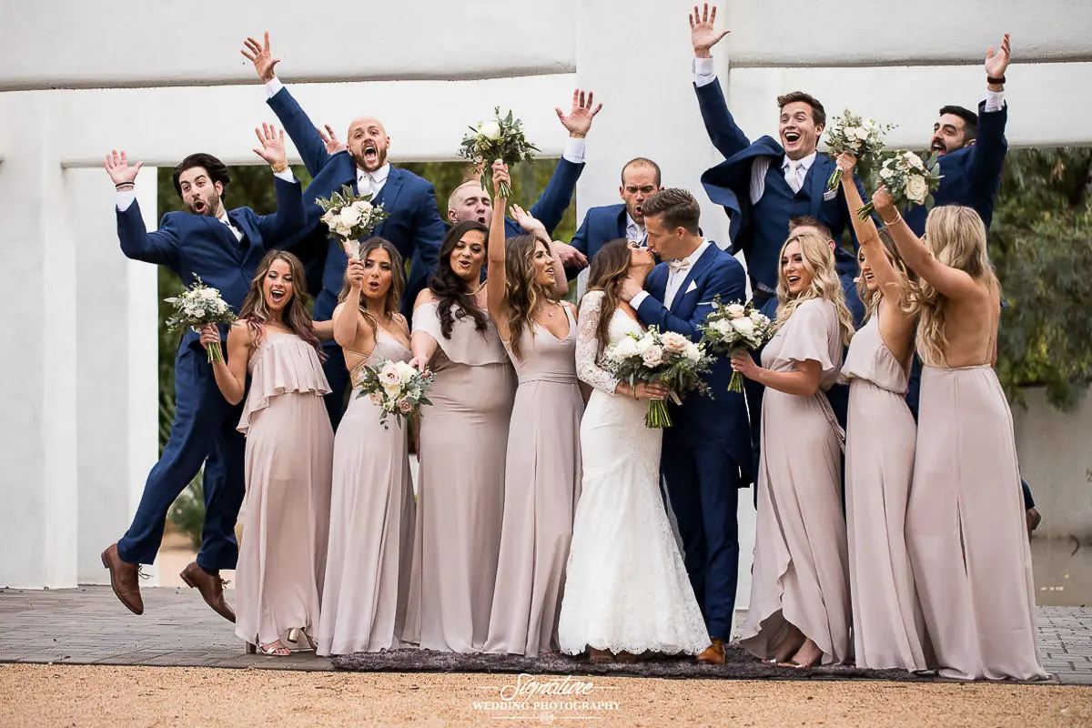 Wedding Party Photos A joyful wedding party celebrates as the bride and groom kiss at the center, captured perfectly by wedding photography. Bridesmaids in beige dresses and groomsmen in blue suits surround them, some holding flowers while others cheer with raised arms against a white wall backdrop.