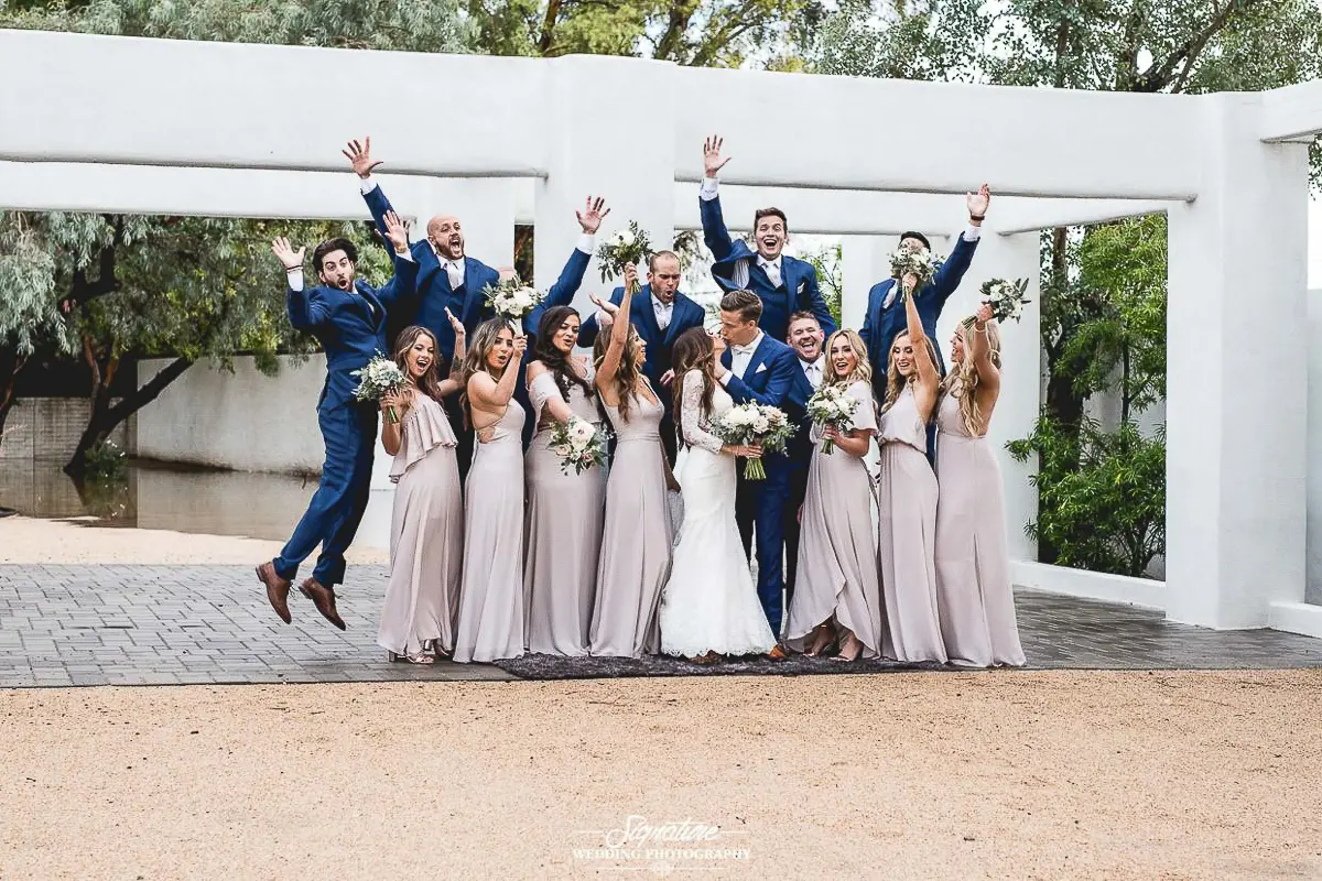 Wedding Party Photos The wedding photography captures a lively party jumping and celebrating outdoors, posed in front of a gleaming white structure with lush greenery. Bridesmaids dazzle in light gray dresses, groomsmen sport stylish blue suits, and the joyous couple beams at the center.