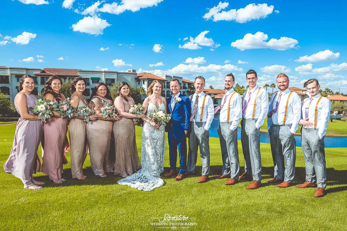 Wedding Party Photos A wedding party poses on a green lawn under a blue sky with clouds, captured through exquisite wedding photography. The group features eight bridesmaids in pastel dresses, a bride in white, a groom in blue, and five groomsmen in gray pants and suspenders.