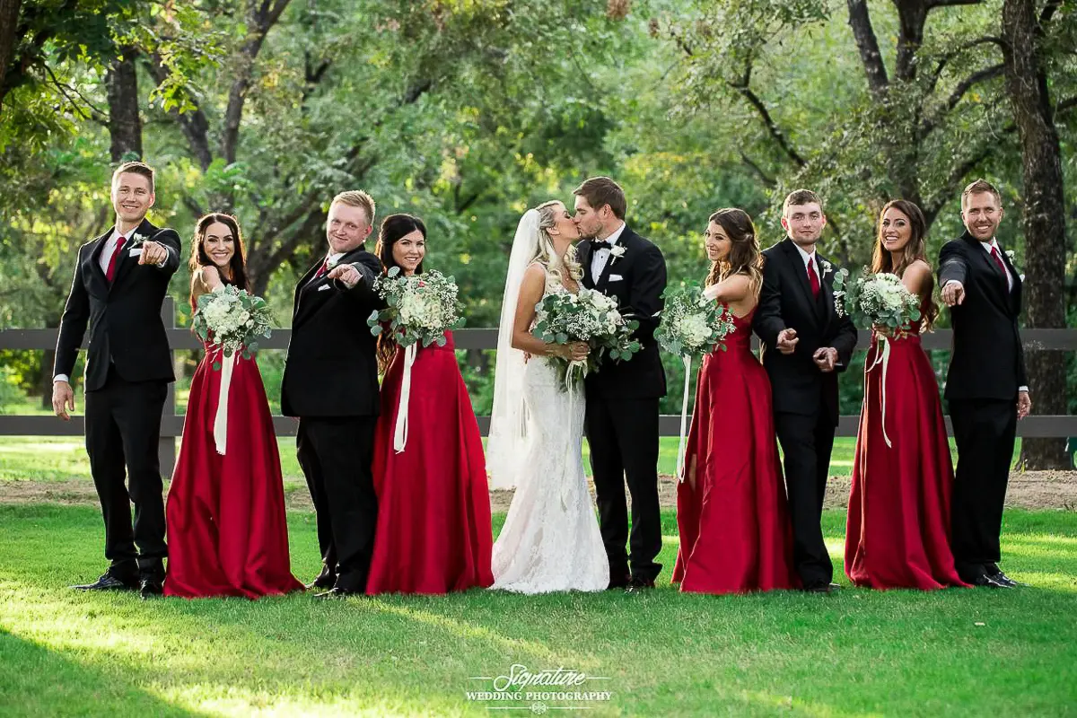 Wedding Party Photos A bride and groom share a kiss outdoors, captured beautifully by wedding photography. Surrounded by a bridal party, bridesmaids in red dresses hold bouquets while groomsmen in black suits point playfully, all set against a backdrop of lush grass and towering trees.