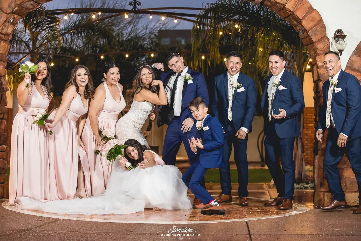 Wedding Party Photos A bride and groom strike playful poses with their wedding party under a decorated archway, capturing the essence of wedding photography. The group includes two children in blue suits and four women in pink dresses, all smiling and displaying energetic gestures amid festive lighting.