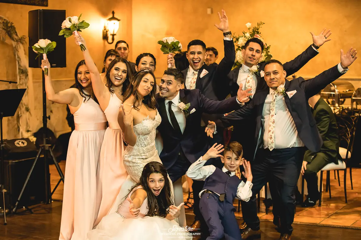 Wedding Party Photos A joyful wedding photography session captures the energetic party indoors. The bride and groom stand in front, surrounded by bridesmaids in pink dresses and groomsmen in suits, while two smiling children crouch happily before them. Everyone looks festive and full of joy.
