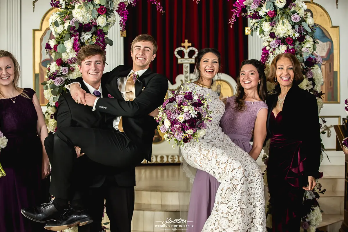 Wedding Party Photos A joyful group stands at a wedding ceremony, captured flawlessly in timeless wedding photography. Two men in suits lift a smiling bride in a white lace dress as bridesmaids in purple dresses and another woman in a dark outfit celebrate. Floral decorations in purple and white adorn the background.