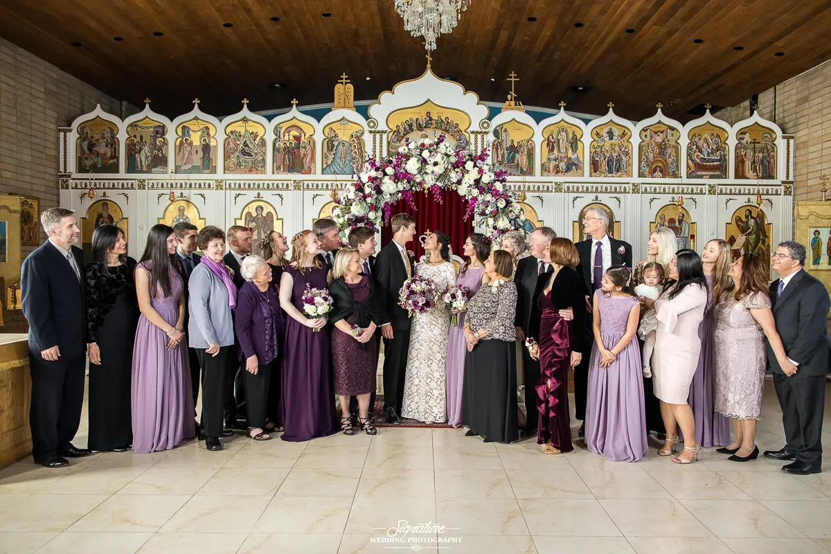 Wedding Party Photos A wedding group photo in a church captures the bride and groom at the center, showcasing exquisite wedding photography. Guests dressed in formal attire, many in shades of purple, surround them. The background features religious iconography and a stunning floral arrangement.