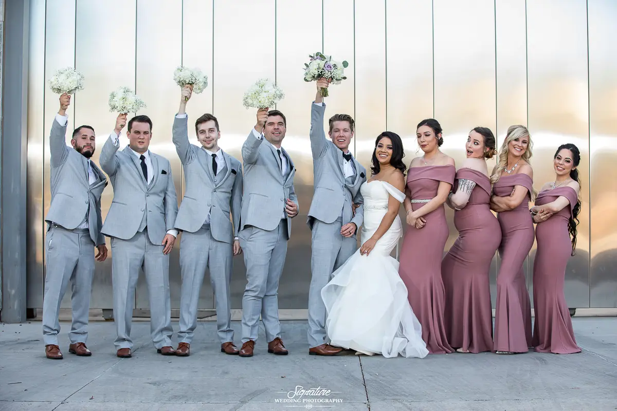 Wedding Party Photos A wedding party poses joyfully against a metallic wall, capturing the essence of wedding photography. Four men in light gray suits stand on the left, raising bouquets. The bride, in a white dress, is flanked by bridesmaids in mauve dresses on the right. Everyone is smiling and celebratory.