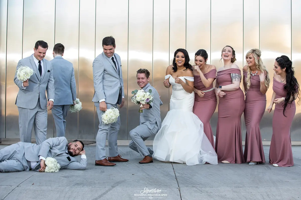 Wedding Party Photos In a delightful display of wedding photography, the bride in a white gown and groom in a light grey suit pose playfully with their lively wedding party. He humorously lies on the ground with a bouquet as bridesmaids in mauve dresses laugh, while groomsmen stand by, some holding flowers.