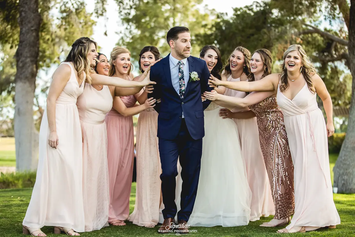 Wedding Party Photos In this charming example of wedding photography, a groom stands playfully in the center, surrounded by eight bridesmaids reaching towards him. They are all smiling and wearing elegant, light-colored dresses. The scene is beautifully set outdoors with trees in the background.
