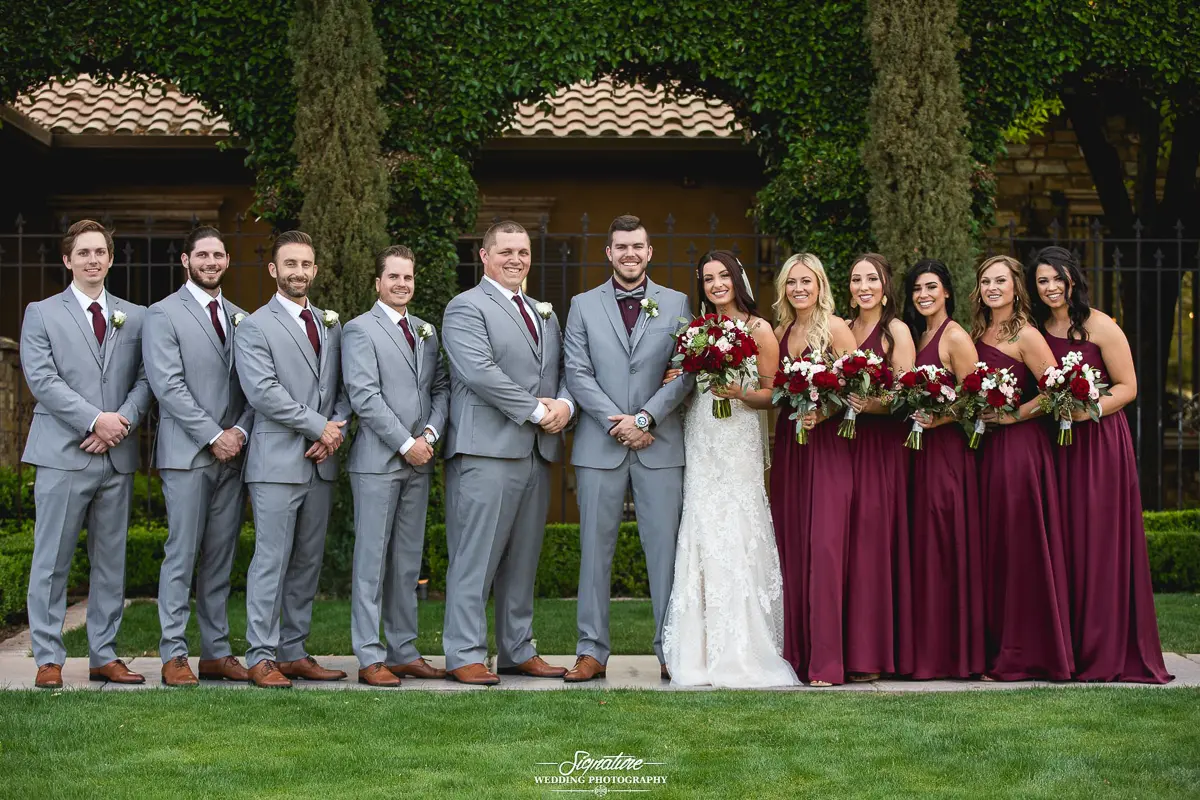 Wedding Party Photos A wedding party poses amidst lush greenery, capturing a perfect moment for wedding photography. Four men in gray suits with burgundy ties stand alongside five women in burgundy dresses. The bride, radiant in white, stands by the groom as they all hold bouquets.