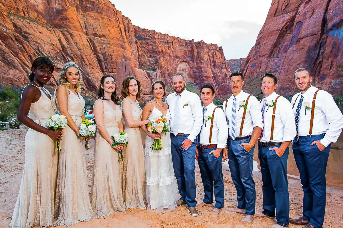 Wedding Party Photos The wedding photography captures the party posing against stunning red rock formations. The bride and groom stand at the center, surrounded by groomsmen in suspenders and bridesmaids in beige dresses, each holding flowers. The scene is vibrant with natural beauty and elegance.