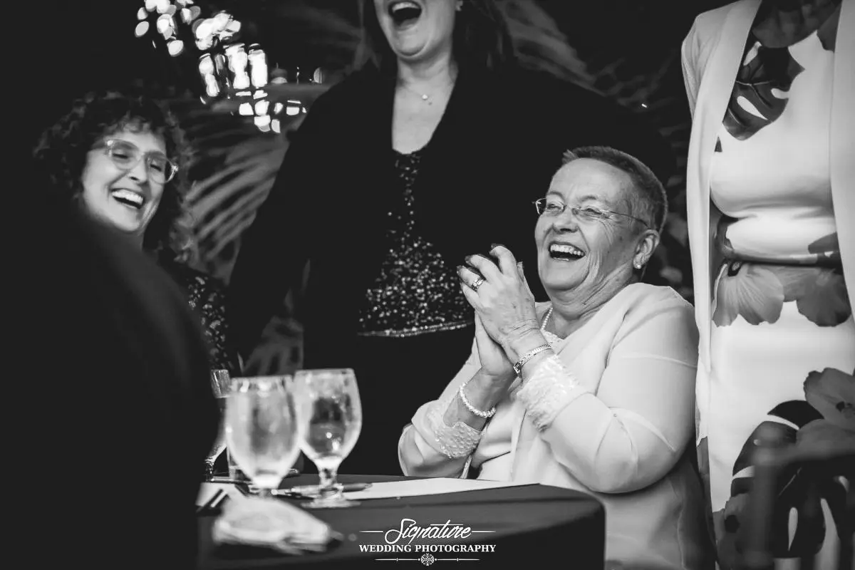 Wedding Photography Candid Moments A group of people gathered around a table, laughing and enjoying themselves at what seems like a wedding photography session. One person in the foreground, wearing glasses, is clapping. Glasses of water and a napkin are on the table. The scene is in black and white.