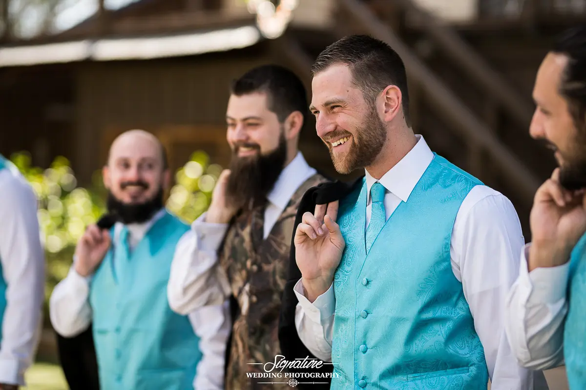 Wedding Photography Candid Moments Groomsmen in turquoise vests stand smiling and laughing outdoors, with one holding his suit jacket over the shoulder. Captured in a sunny location, they exude relaxed joy—a perfect moment of wedding photography magic.