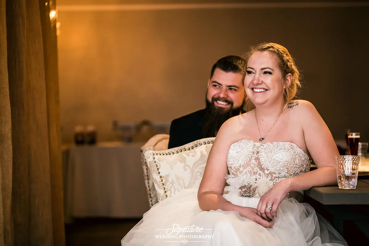 Wedding Photography Candid Moments A bride in a white strapless wedding dress sits smiling next to her bearded groom, partially obscured. The soft lighting and beige backdrop capture the intimate moment in this stunning piece of wedding photography. A glass of water rests on the table before them.