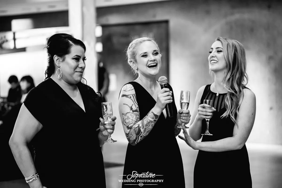 Wedding Photography Candid Moments Three women in black dresses hold champagne glasses at what seems like a lively wedding event. The woman in the center, adorned with tattoos and holding a microphone, is speaking or singing. They are indoors, smiling radiantly as they celebrate this joyous occasion.