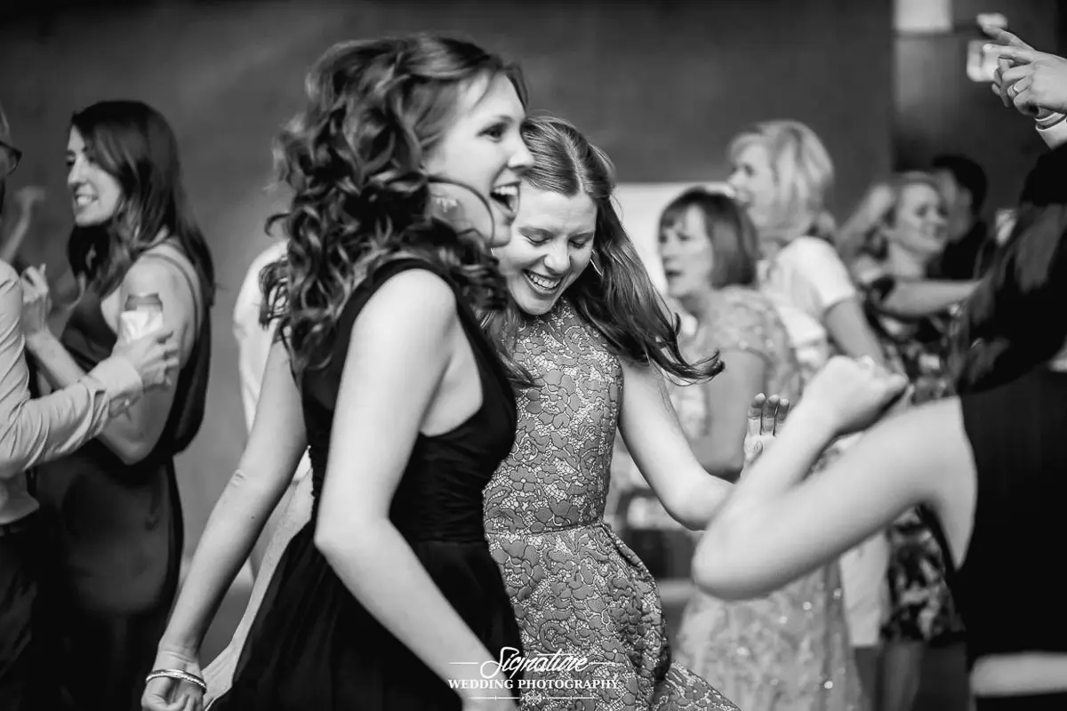 Wedding Photography Candid Moments Black and white photo of two women joyfully dancing at a wedding, surrounded by others reveling in the moment. Laughter fills the air as high spirits abound. In the background, guests engage in cheerful conversation, capturing the essence of wedding photography bliss.