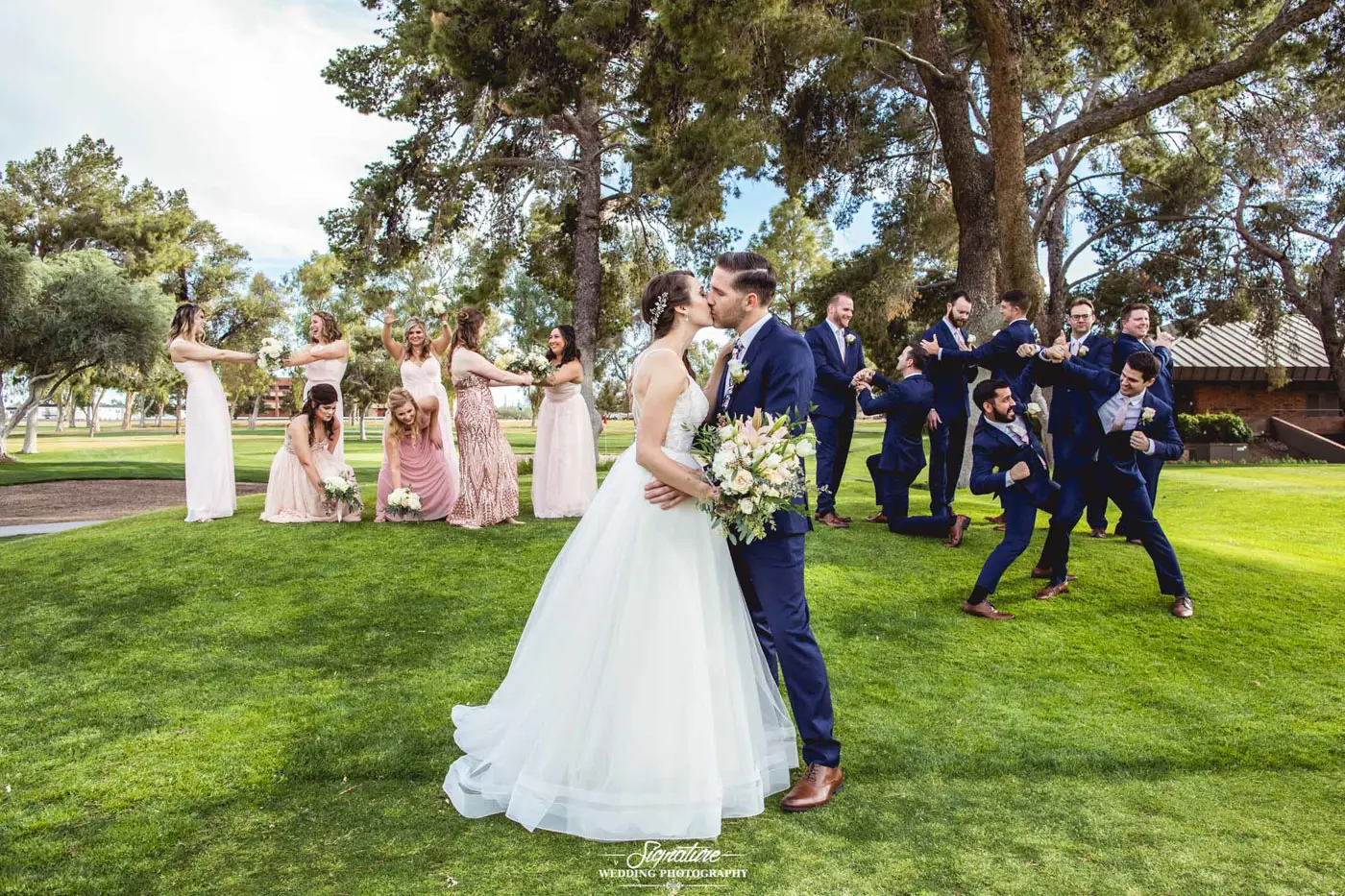 Bride and groom kissing with wedding party in funny poses behind
