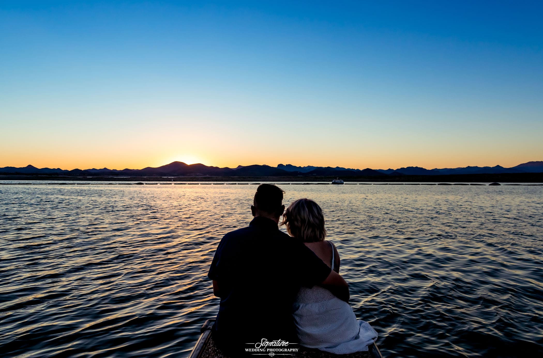 Man's arm around woman sitting at lake during sunset