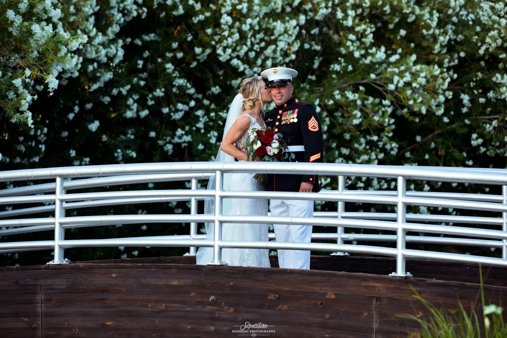 Bride kissing groom in uniform on the cheek on bridge