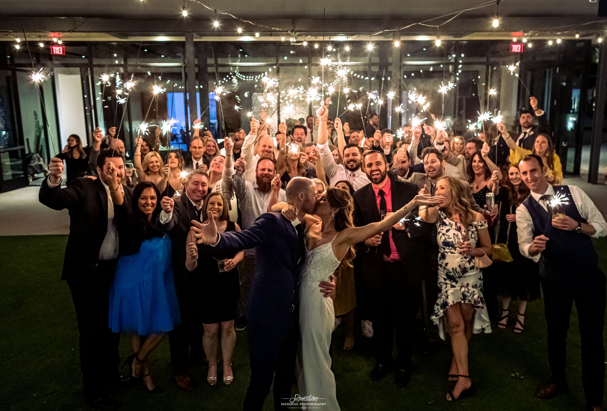 Bride and groom kiss in front of wedding guests with sparkers group shot
