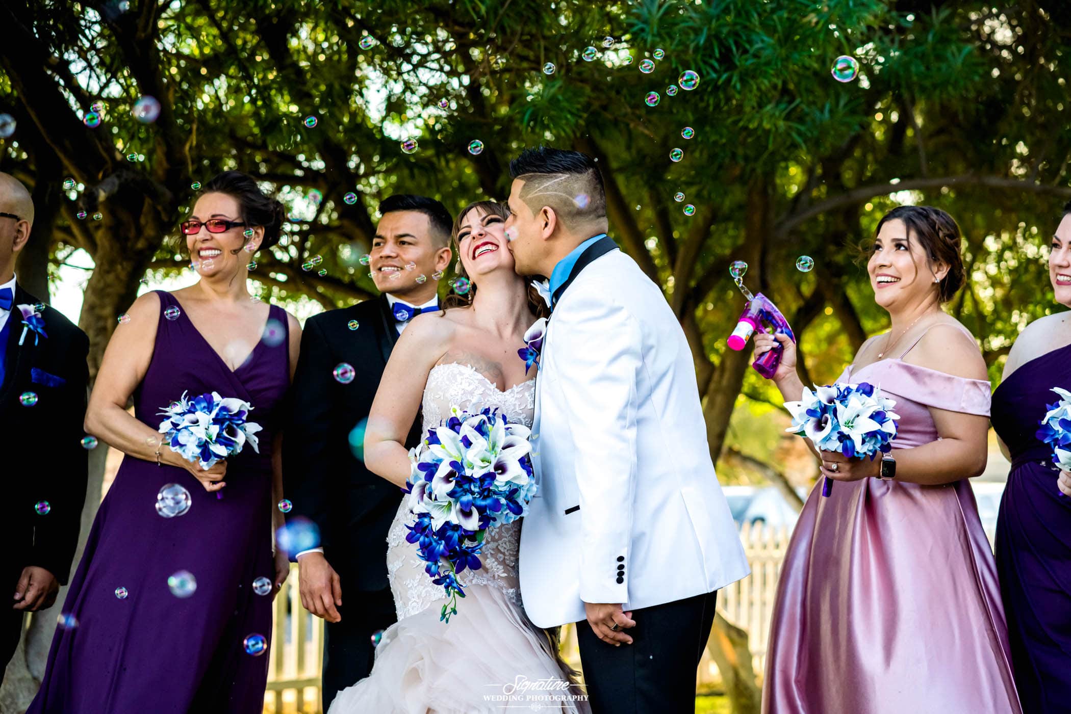 Groom kissing bride on cheek with wedding party and bubbles