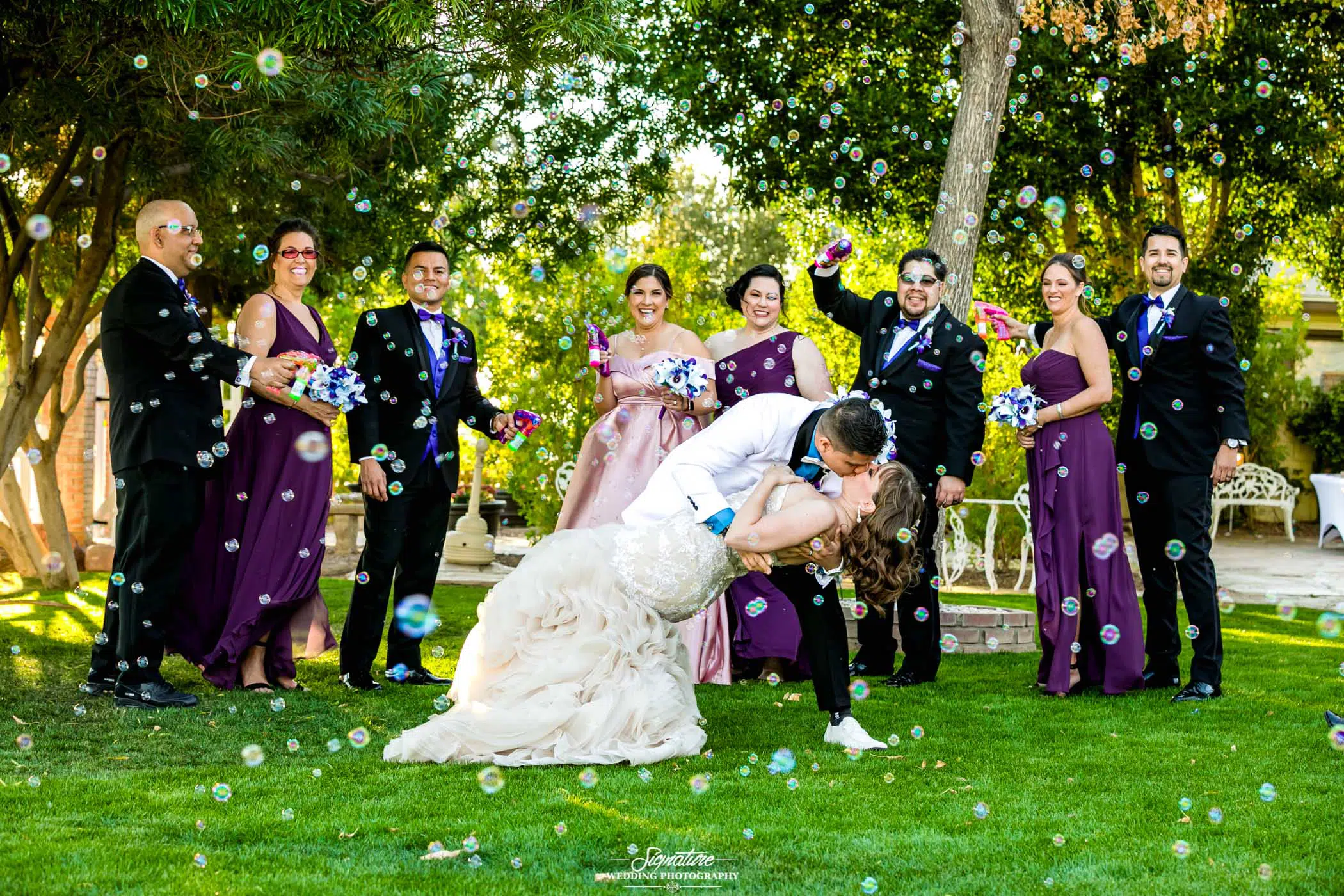 Groom dipping and kissing bride in front of wedding party with bubbles