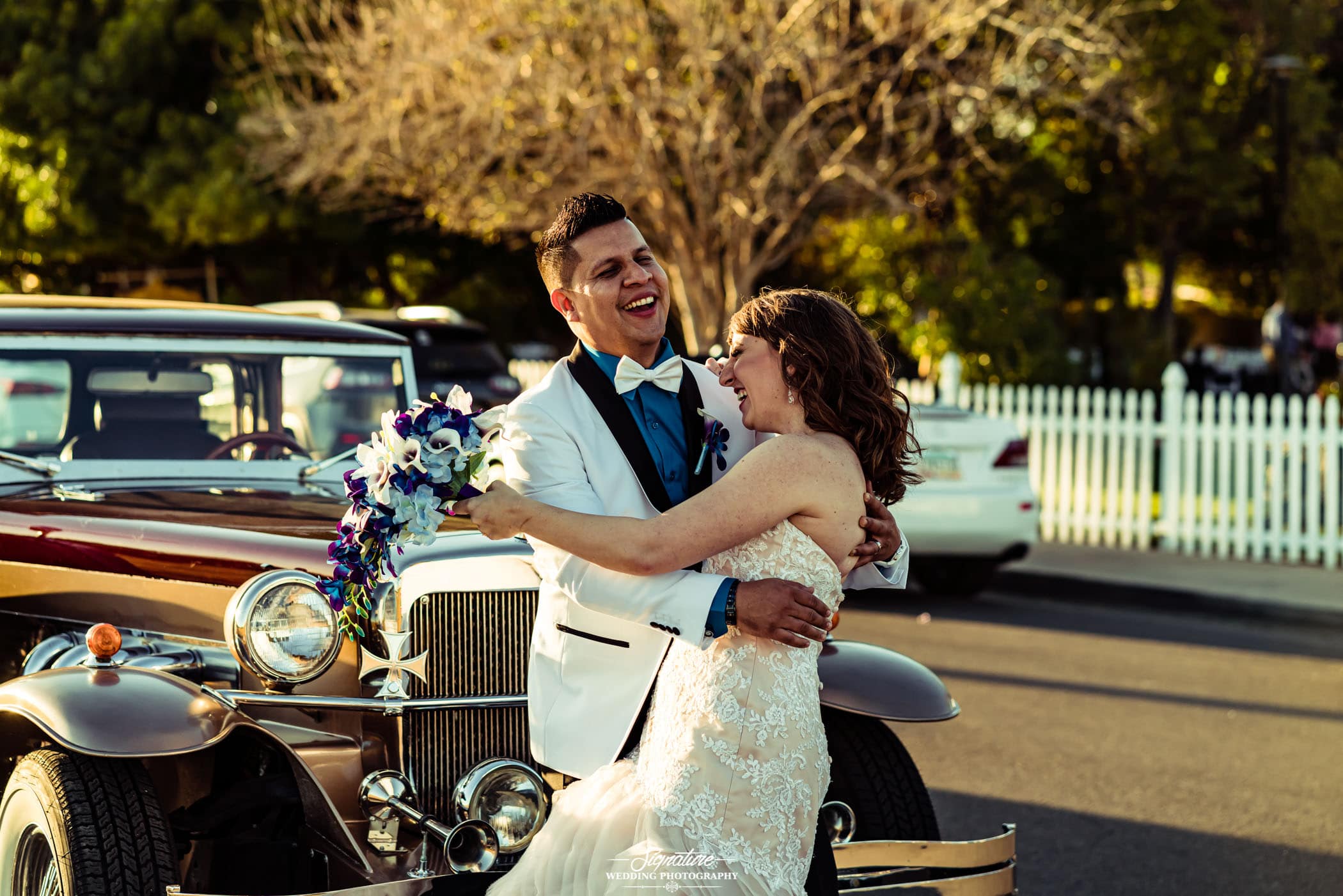 Bride and groom smiling and hugging in front of vintage car