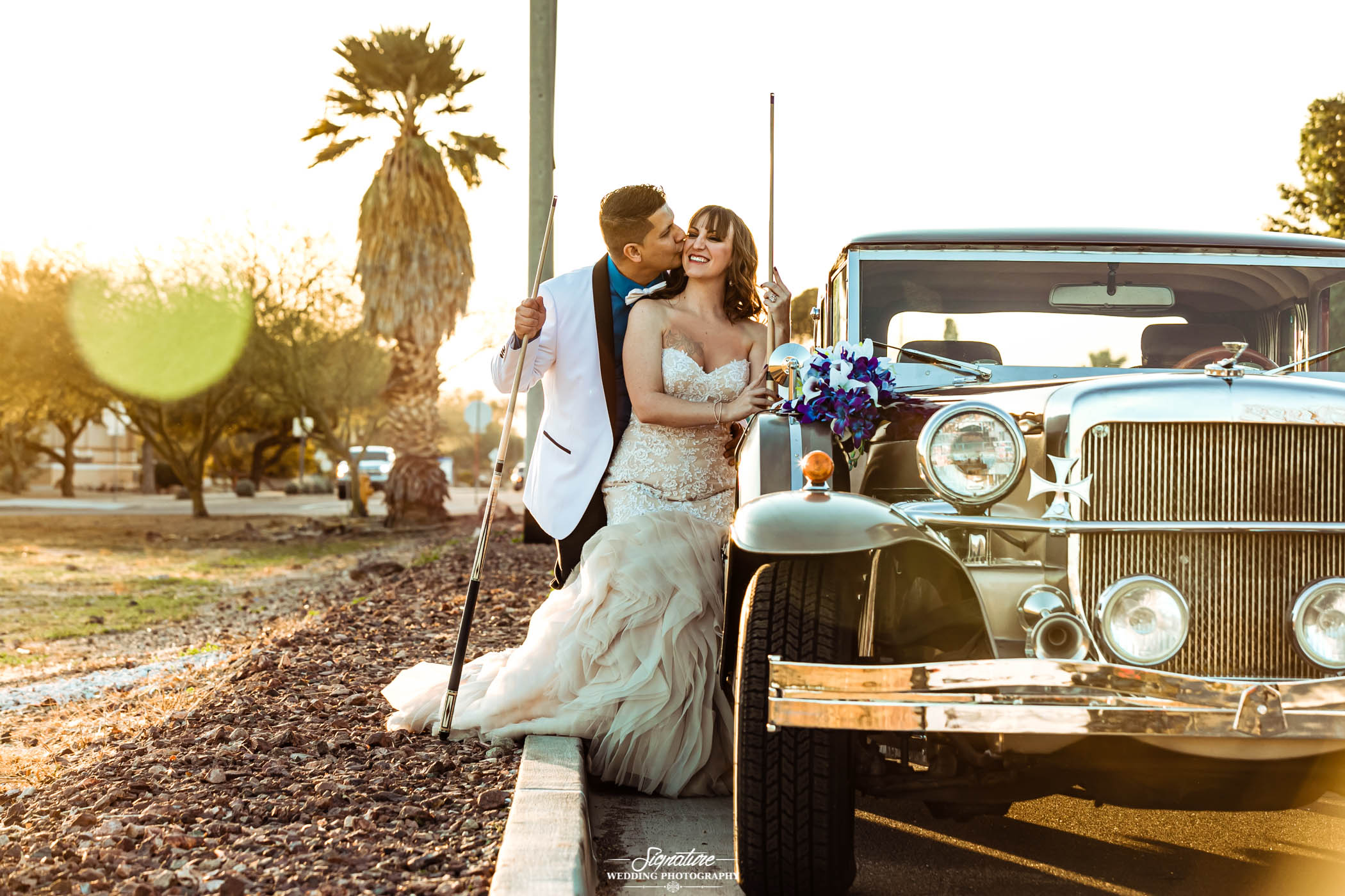 Groom kissing bride on cheek outside vintage car with pool sticks