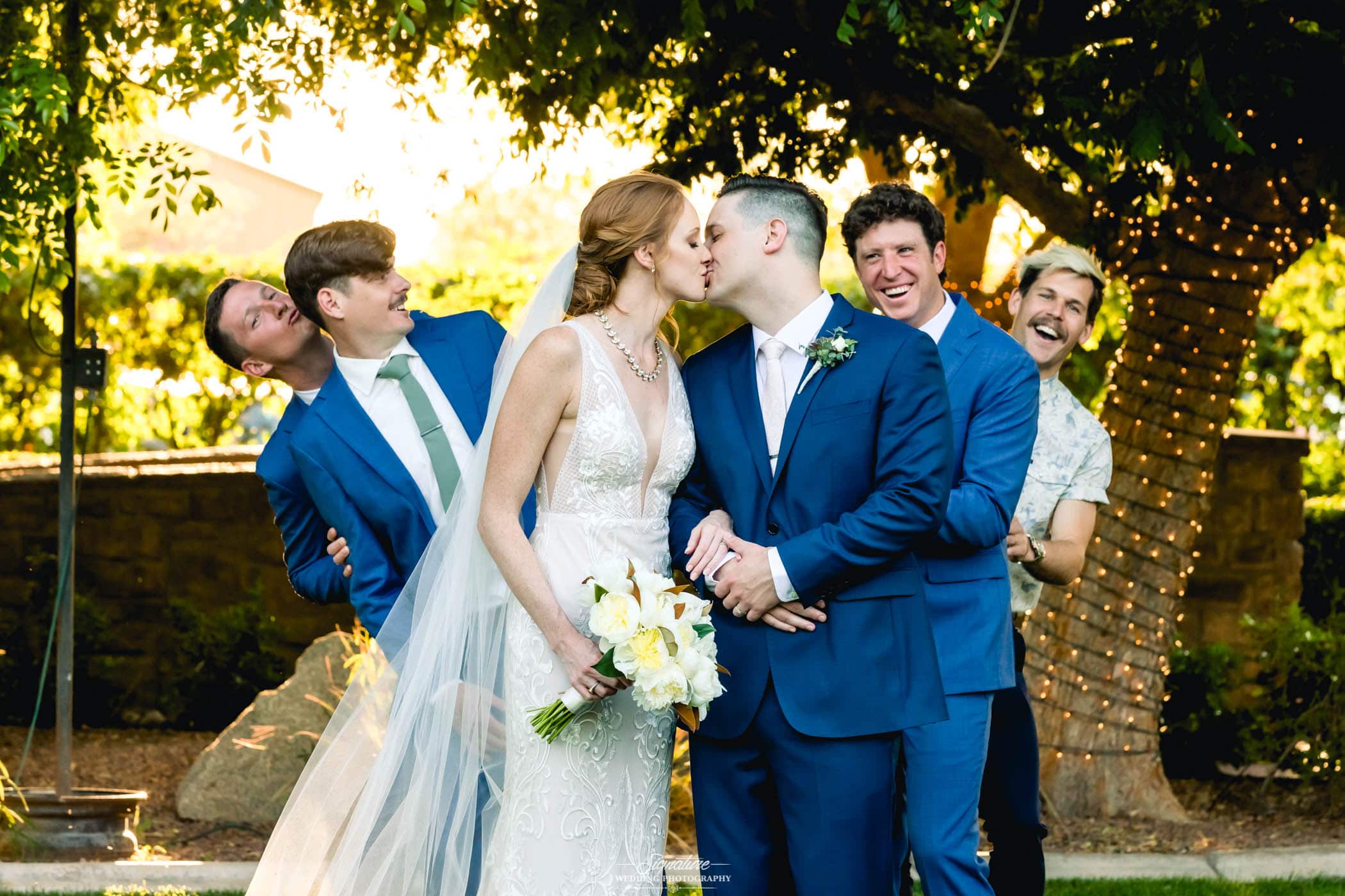 Bride and groom kiss with groomsmen behind reacting
