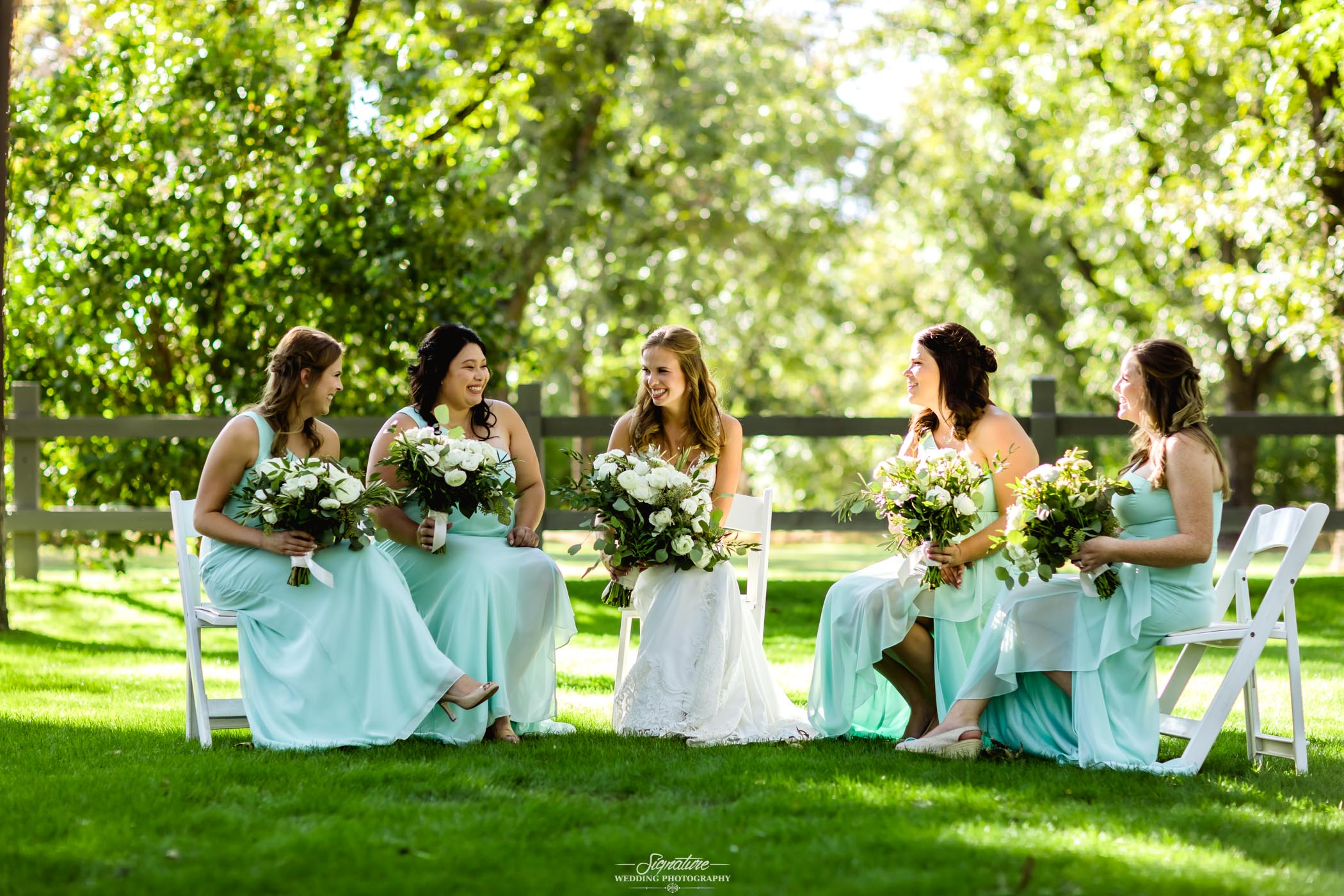 Bride and bridesmaids sitting on chairs outside smiling