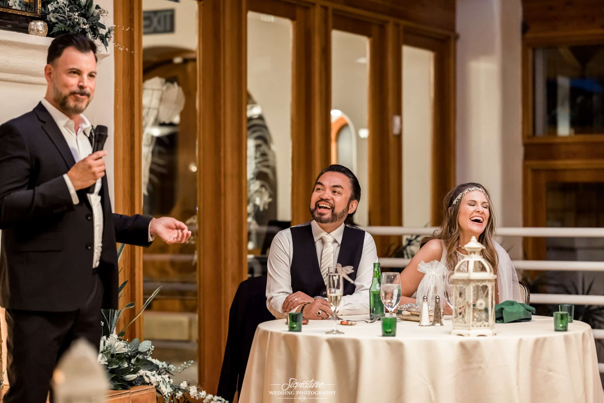 Bride and groom laugh while man gives speech at wedding reception