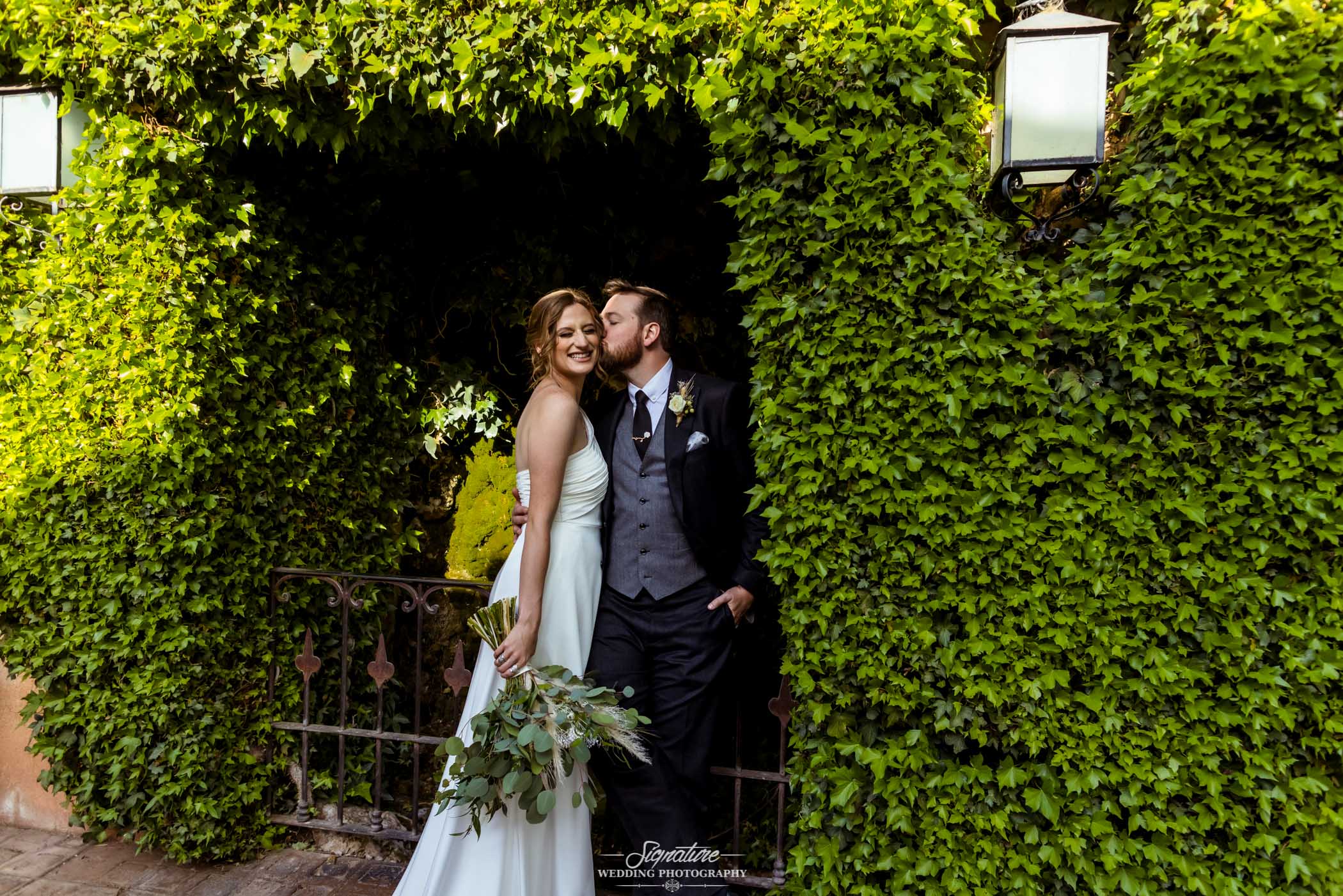 Groom kissing bride on cheek in vine archway
