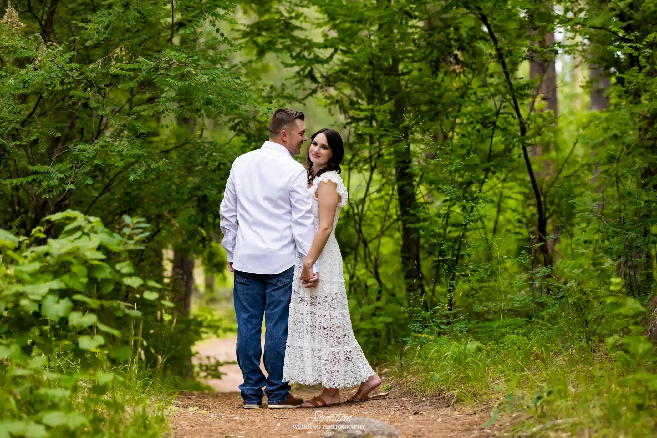 Image by Signature Wedding Photography engagement photo of a couple on a trail, holding hands with the fiancée looking back towards the camera
