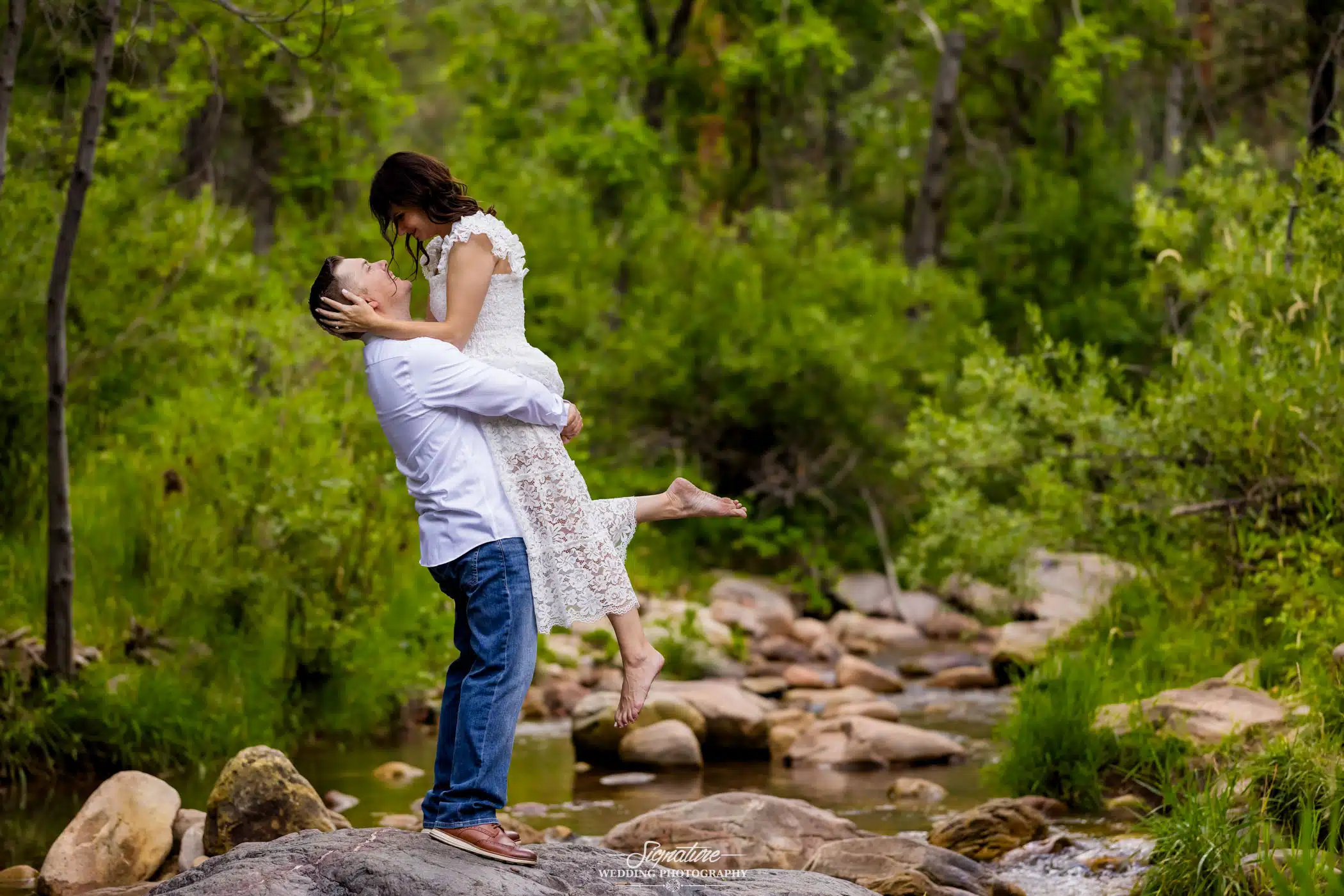 Image by Signature Wedding Photography man holding his fiancée up in the woods by a creek