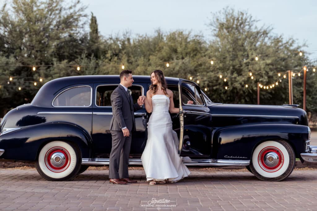 bride and groom standing by the side of a vintage car looking at each other