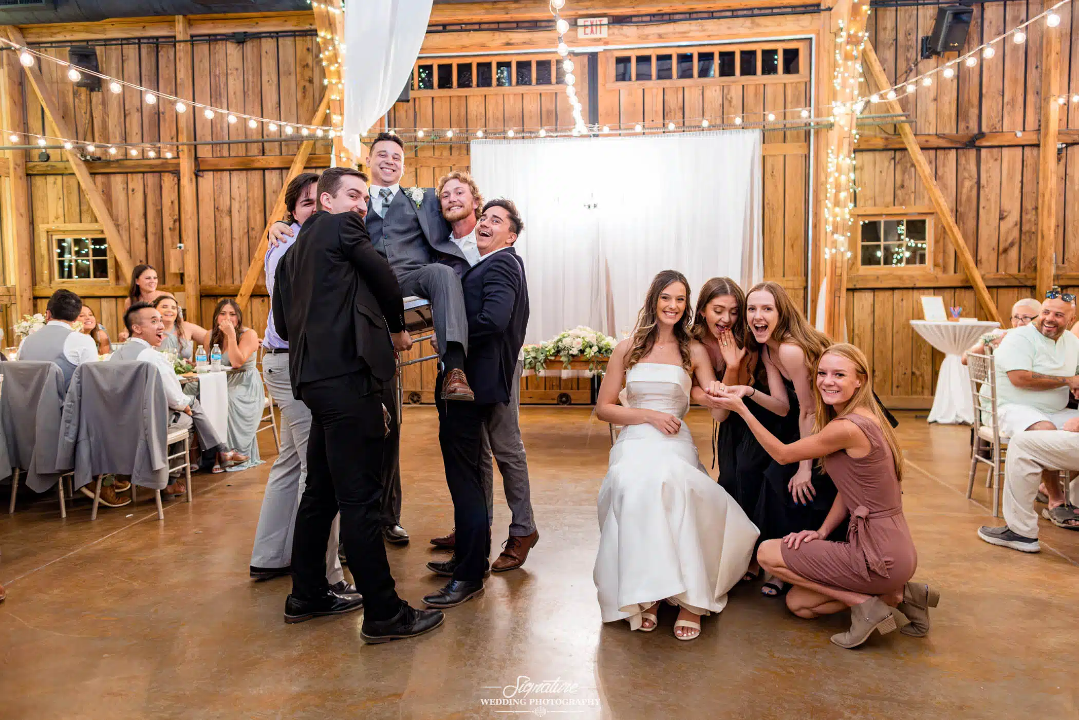 Image by Signature Wedding Photography wedding party photo groomsmen holding up groom in chair, bridesmaids surrounding bride