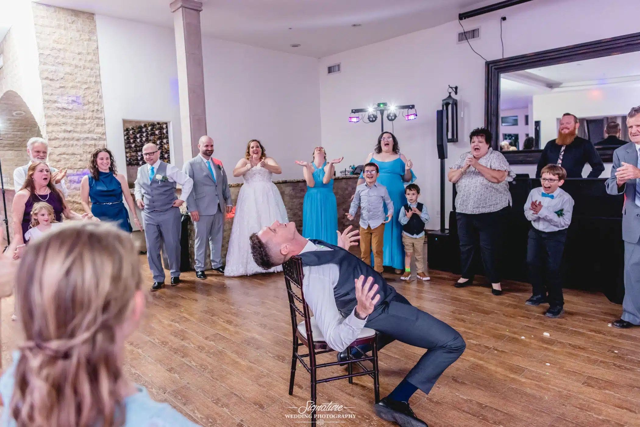 Image by Signature Wedding Photography groomsman sitting awkwardly in a chair to the delight of those watching his antics