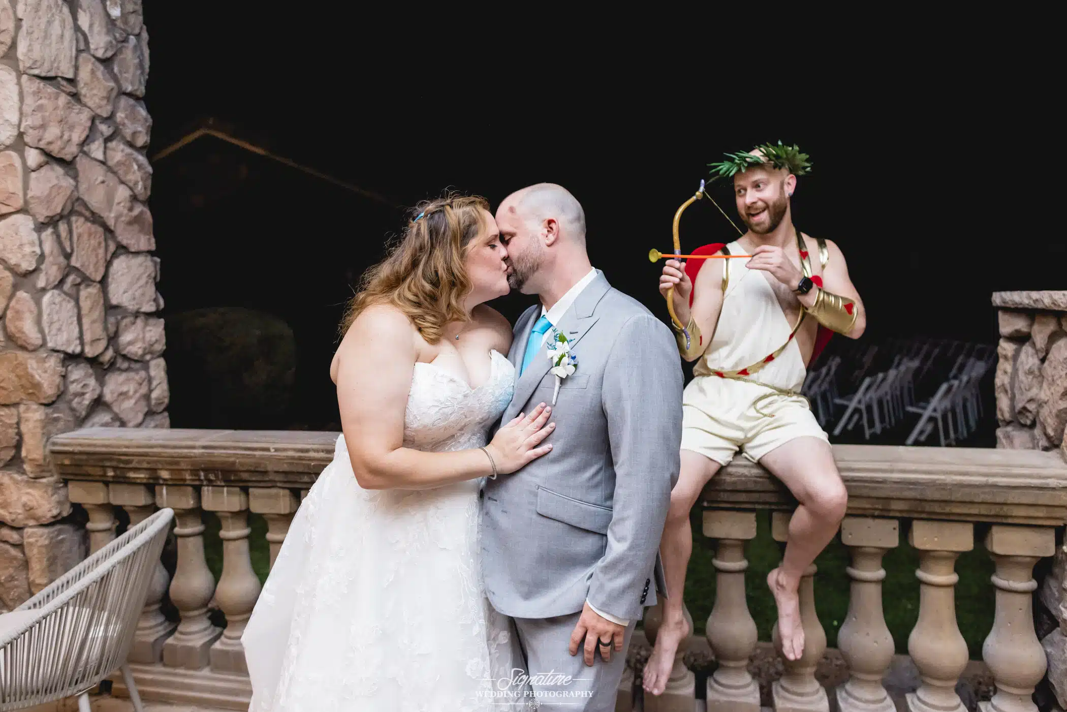 Image by Signature Wedding Photography bride and groom kissing while a Cupid behind them points his arrow at them