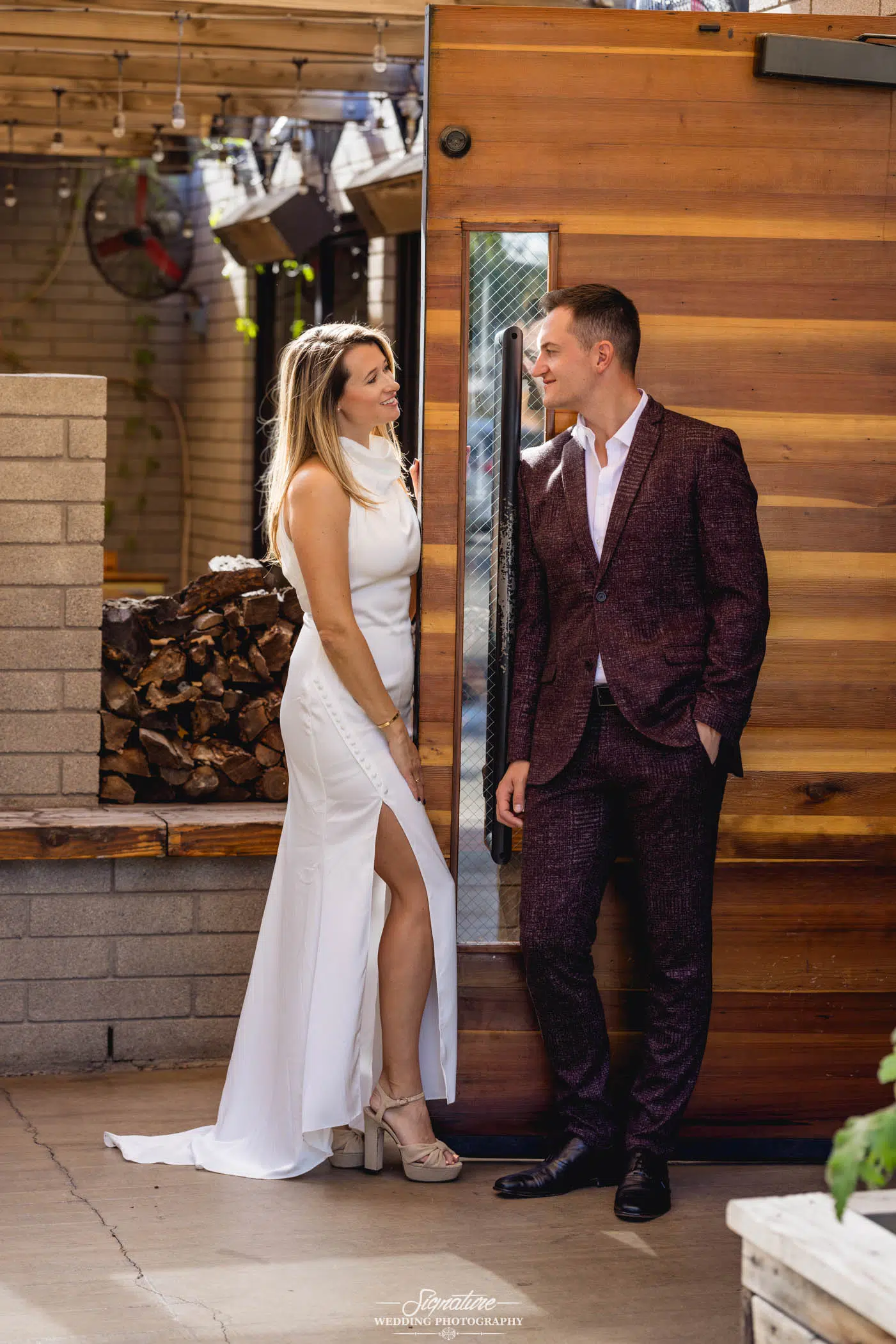 Image by Signature Wedding Photography A woman in a white dress and a man in a burgundy suit stand smiling at each other near a wooden wall, capturing the perfect moment for wedding photography. The woman leans against the wall, with a stack of firewood nearby, set amidst lush greenery.