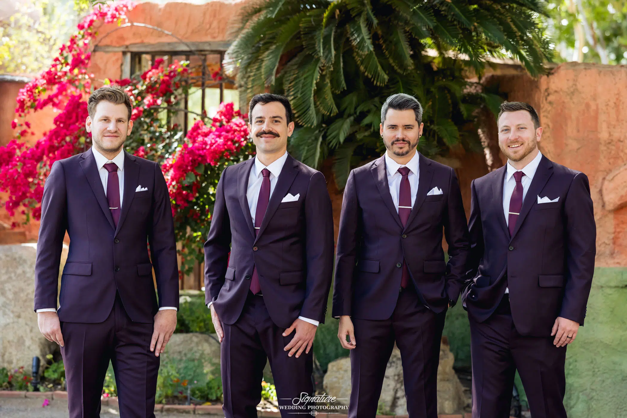 Image by Signature Wedding Photography Four men in dark suits and ties stand outdoors, smiling, as if captured in perfect wedding photography, against a backdrop of vibrant flowers and greenery.