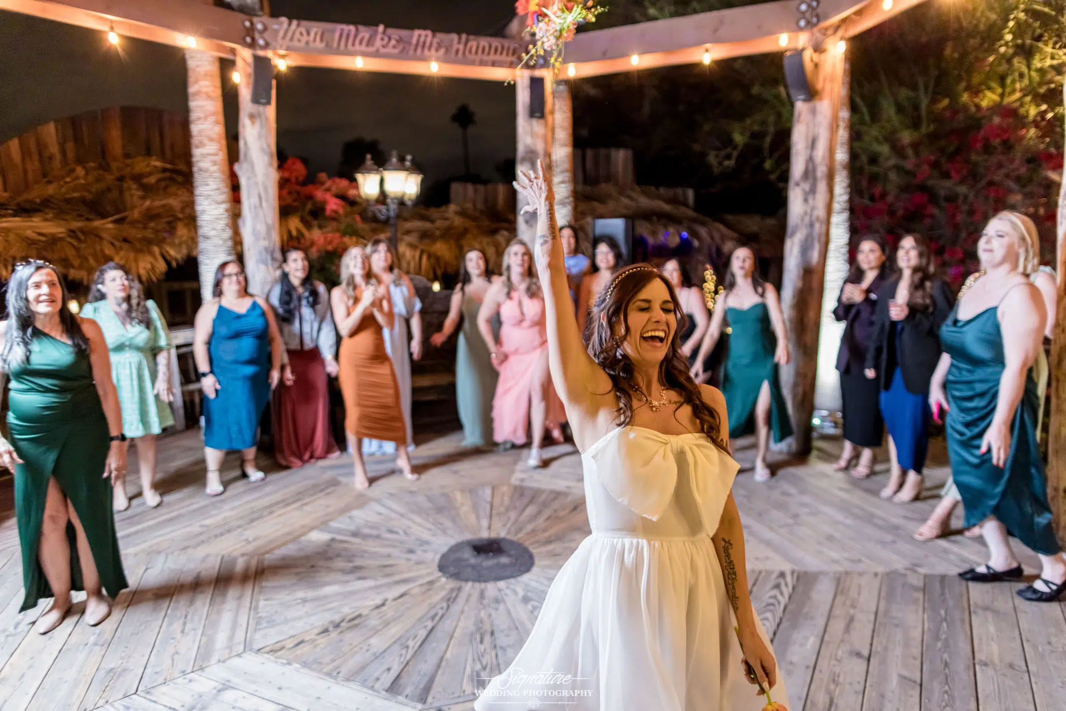 Image by Signature Wedding Photography A bride joyfully holds a bouquet aloft, ready to toss it to a group of eager women behind her. Captured in exquisite wedding photography style, they stand on a wooden deck with tiki-style decor. Lush greenery and warm lights create a festive, tropical atmosphere.
