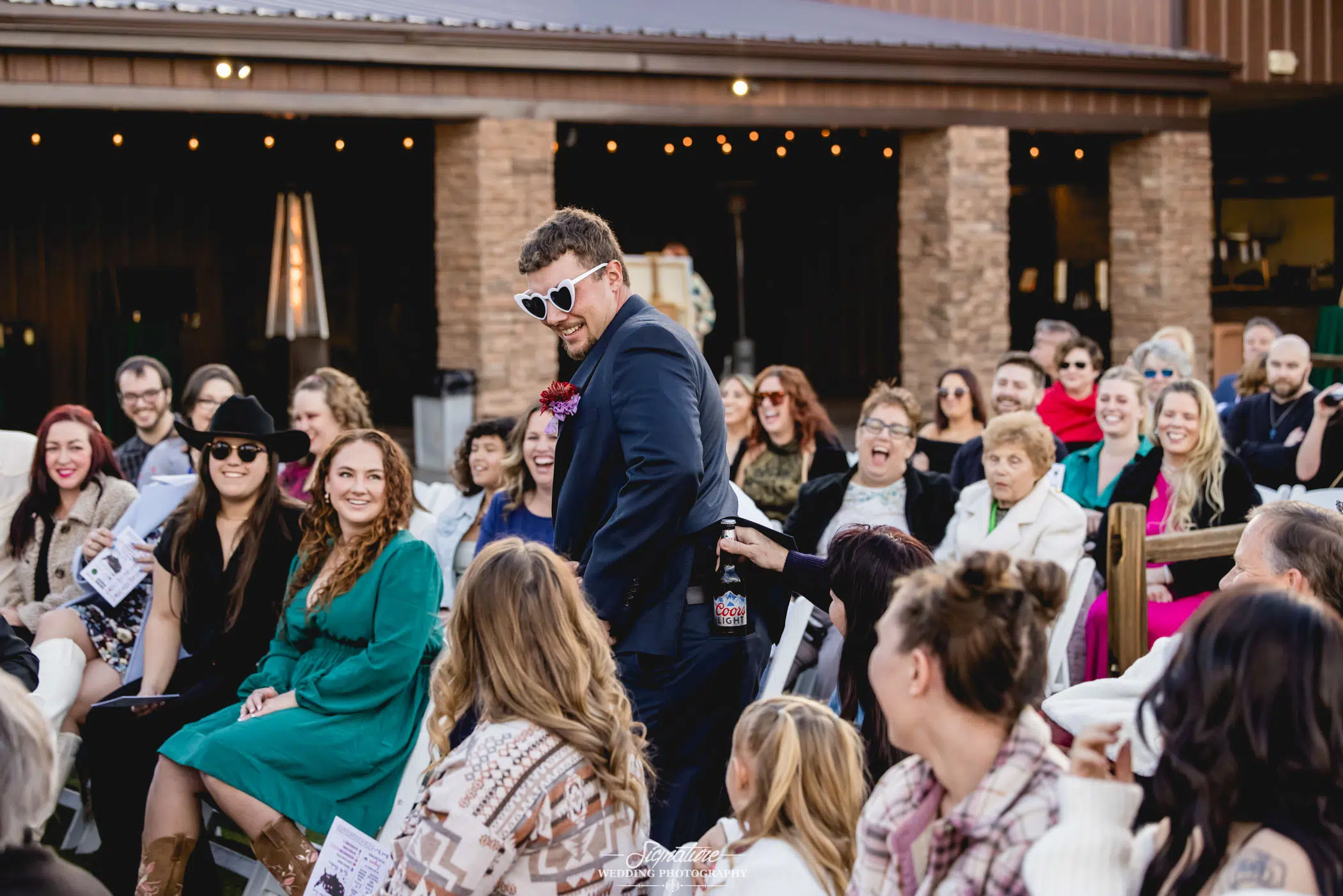 Image by Signature Wedding Photography A man in a dark suit and sunglasses confidently walks down an aisle outdoors, attended by smiling guests capturing the moment. The festive scene is perfect for wedding photography, with a rustic building and string lights adding to the charm.