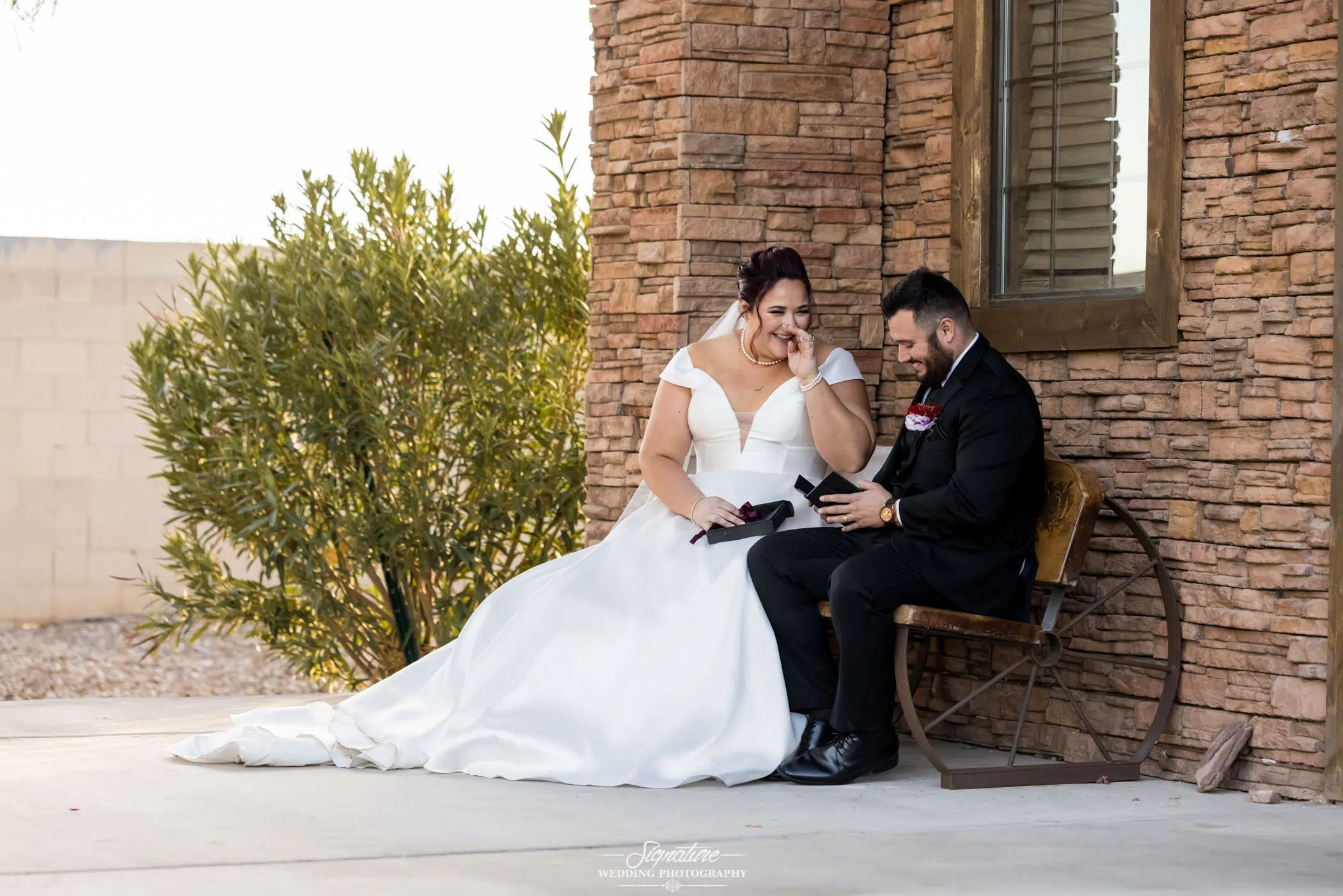 Image by Signature Wedding Photography A bride and groom are sitting on a wooden bench beside a stone wall. The bride, in a white dress, is smiling, while the groom, in a black suit, shows her something. This wedding photography captures them looking joyful and relaxed.
