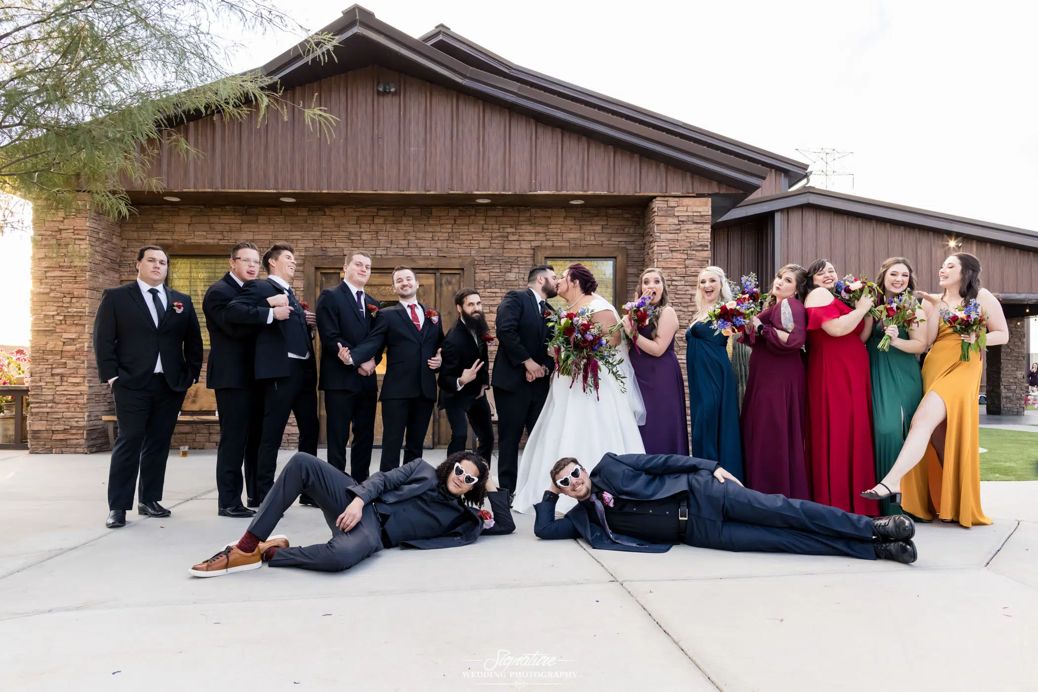 Image by Signature Wedding Photography A wedding party poses playfully outside a rustic building, capturing the essence of wedding photography. The bride and groom kiss in the center, surrounded by attendants in formal attire, each in different colored dresses or suits. Two groomsmen lie on the ground in front, striking relaxed poses.