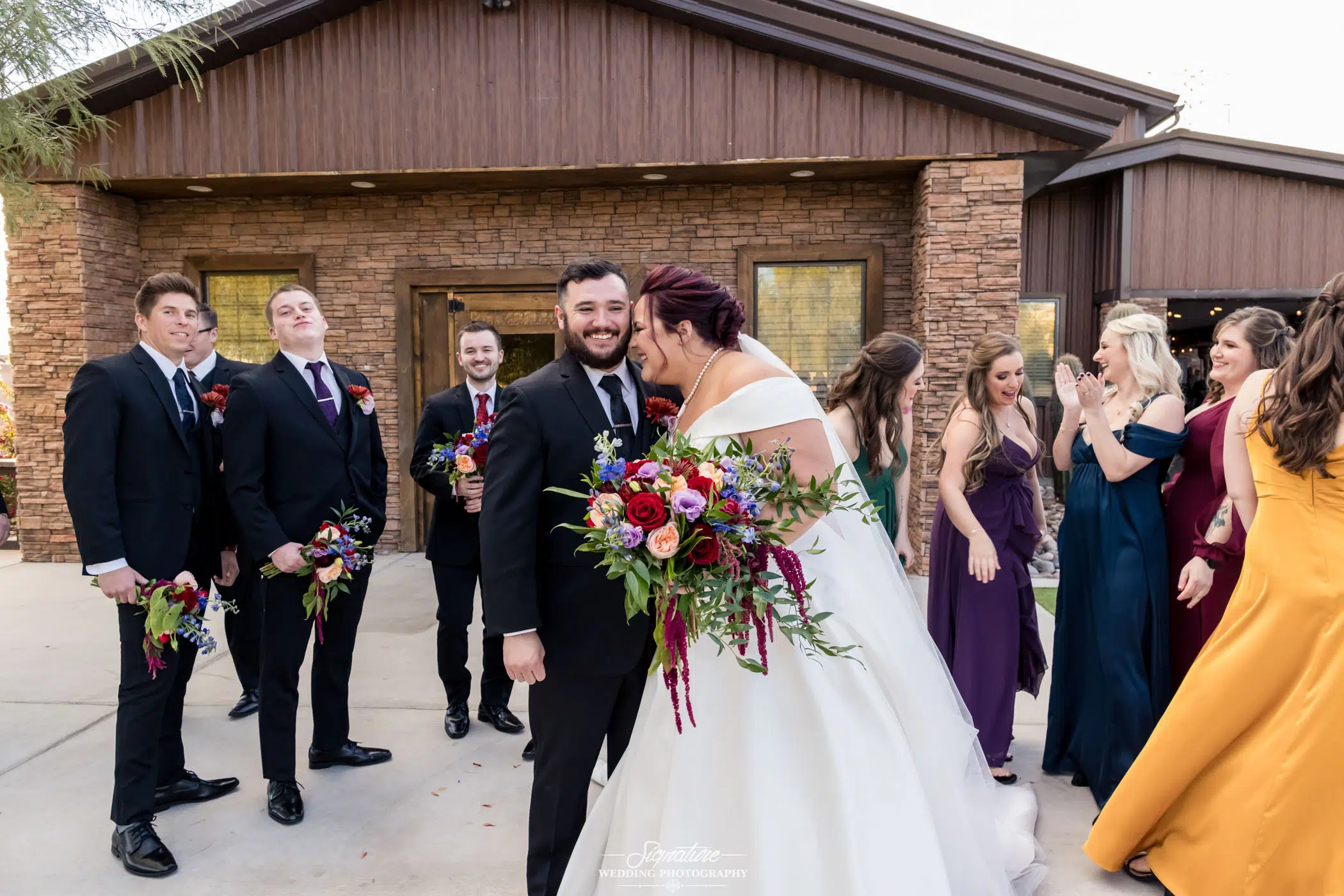 Image by Signature Wedding Photography A wedding couple stands joyfully outside a rustic building, captured beautifully through expert wedding photography. The bride, in a white gown, holds a vibrant bouquet. The groom, in a black suit, smiles as bridesmaids in various dresses and groomsmen in suits pose happily beside them.