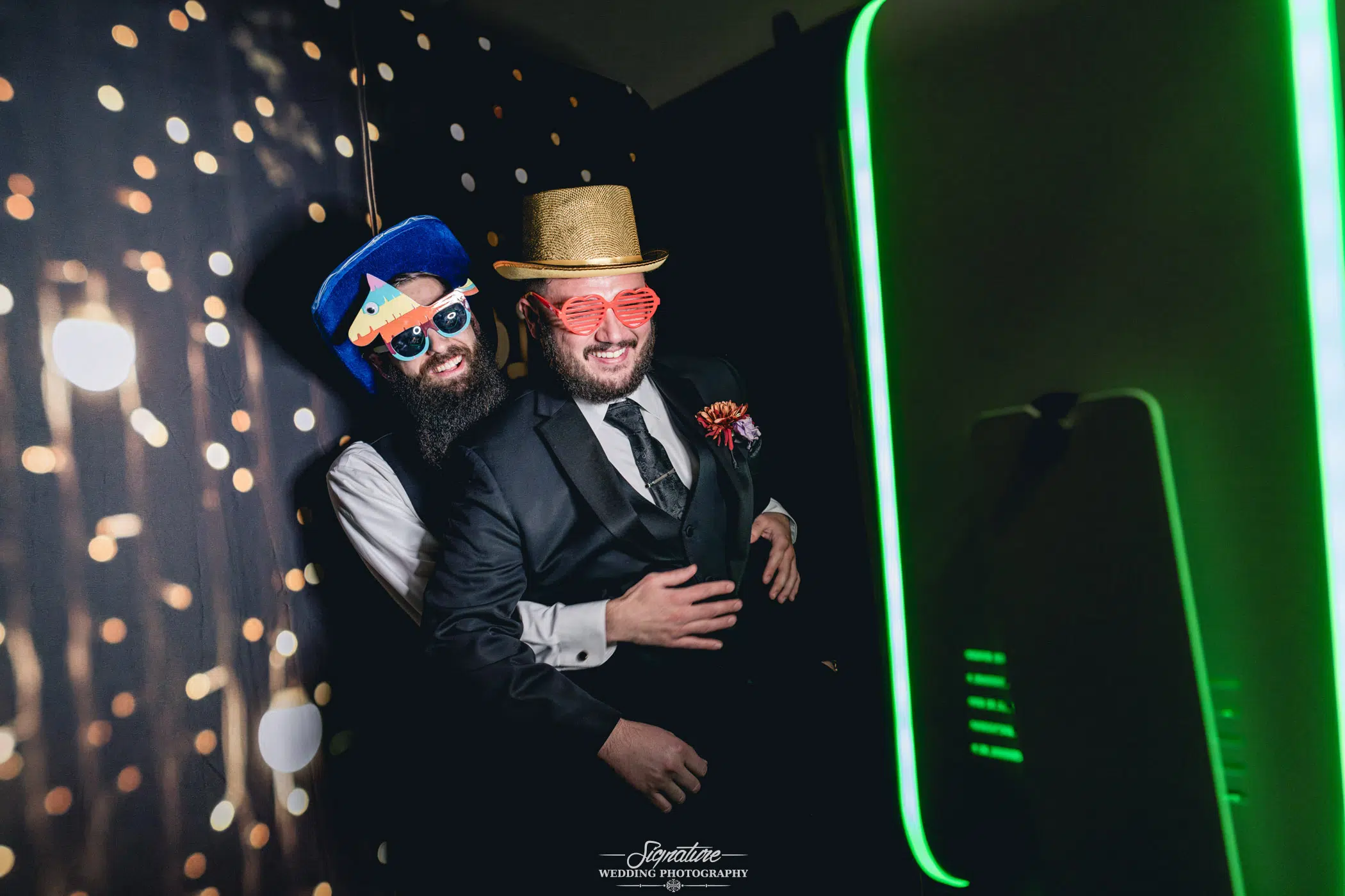 Image by Signature Wedding Photography Two men in suits pose playfully in a photo booth at a wedding. One wears a colorful hat with a propeller and large red sunglasses; the other sports a gold glittery hat. Both smile, standing against bokeh lights beside the bright photo booth screen, adding fun to the wedding photography session.