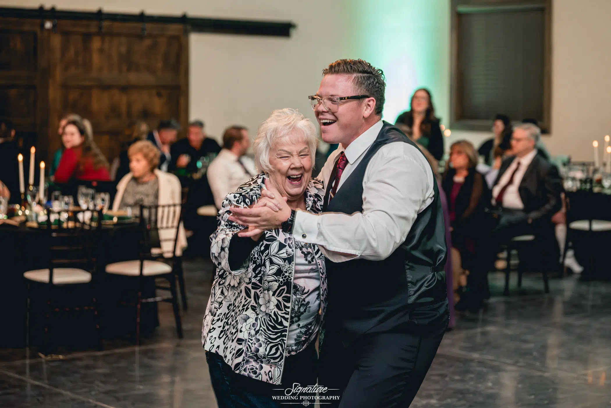 Image by Signature Wedding Photography A man in a vest and tie dances joyfully with an elderly woman in a patterned jacket at a lively indoor wedding photography event. They are surrounded by seated guests at tables, some watching and smiling.