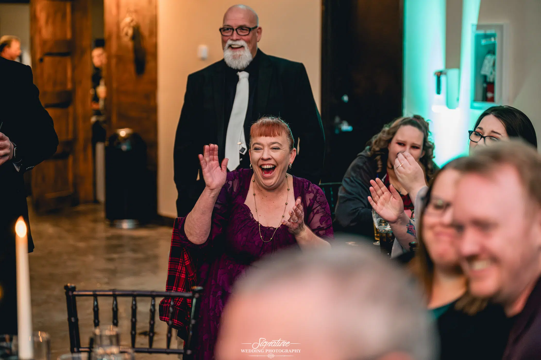 Image by Signature Wedding Photography A woman in a purple dress claps and smiles enthusiastically at an indoor wedding photography event. People around her join in the cheerful applause, enjoying the moment. A man in a suit with a beard stands smiling contentedly in the background.