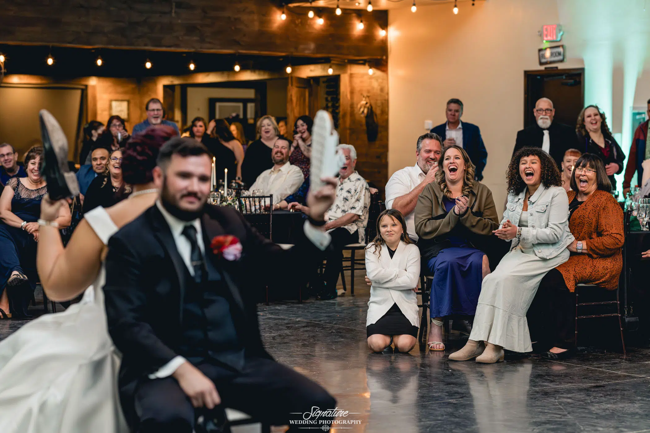 Image by Signature Wedding Photography A couple sits back-to-back on chairs, each lifting a shoe in the air—a delightful moment of wedding photography. The bride, in her white dress, and the groom, suited up, are cheered by guests who laugh and smile under warmly glowing string lights.